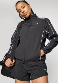Jovem mulher com um casaco desportivo preto da Puma e calções, a carregar um saco de ginásio preto sobre o ombro, posando com confiança.