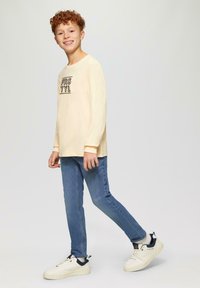 Helles beiges Langarmshirt mit schwarzem Text, kombiniert mit blauen Slim-Fit-Jeans und weißen Sneakers mit marineblauen Akzenten.