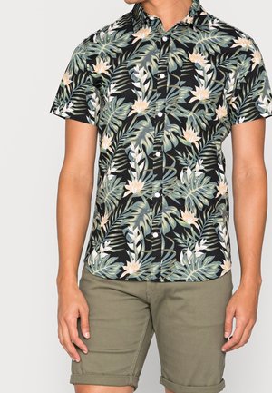 Uomo che indossa una camicia nera a maniche corte con bottoni con motivo a foglie tropicali verdi e beige e pantaloncini verde oliva risvoltati.