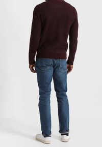 Burgunderfarbener strukturierter Strickpullover, schmal geschnittene blaue Jeans, weiße Sneakers; sichtbare Nähte und Gesäßtaschen an den Jeans.