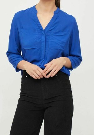 Kobieta o brązowych włosach, ubrana w niebieską bluzkę z długim rękawem i kieszeniami na piersi oraz czarne spodnie z wysokim stanem, ręce złożone na wysokości talii.