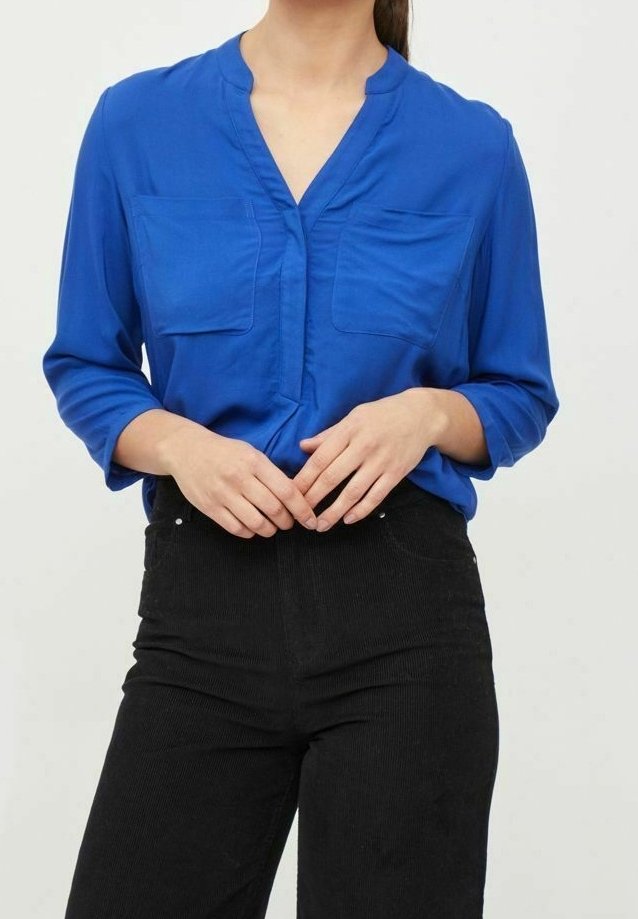 Femme aux cheveux bruns portant un chemisier bleu à manches longues avec des poches poitrine et un pantalon noir taille haute, mains jointes à la taille.