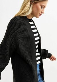 Zwarte gebreide cardigan met open voorkant, textuurstof en lange mouwen, gedragen over een zwart-wit gestreept T-shirt.
