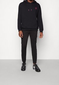 Svart hoodie med en rosa logotyp, tillsammans med svarta cargobyxor med sidofickor. Avslutad med svarta sneakers med vita detaljer.