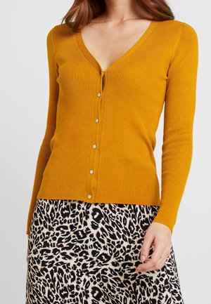 Kobieta w musztardowożółtym prążkowanym kardiganie na guziki i spódnicy w panterkę, ręka spoczywa blisko biodra.