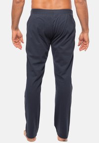 Man staat blootvoets met zijn rug naar de kijker, gekleed in marineblauwe elastische broek met zakken met rits aan de zijkant, tegen een effen witte achtergrond.