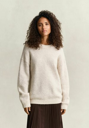 Vrouw met krullend haar, gekleed in een crèmekleurige gebreide trui en een donkerbruine geplooide rok, staand tegen een effen achtergrond.