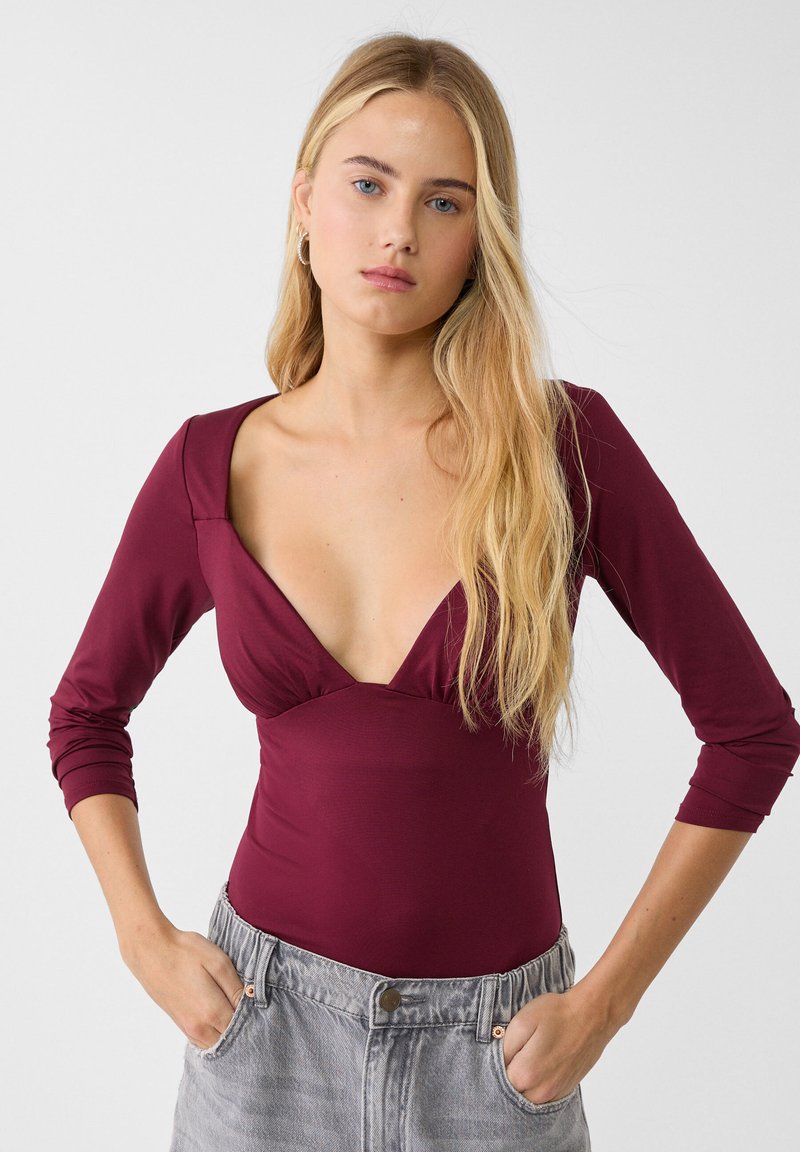 Body bordeaux scollato con maniche lunghe, tessuto morbido, design aderente e dettaglio arricciato sul seno; abbinato a pantaloni corti grigi a vita alta.