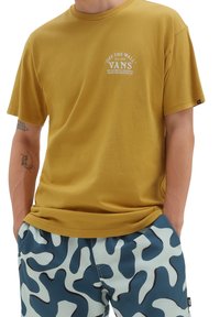 Persona vistiendo una camiseta amarilla mostaza de Vans con logo y pantalones cortos abstractos en azul y blanco, con las manos en los bolsillos y tatuajes visibles en los brazos.