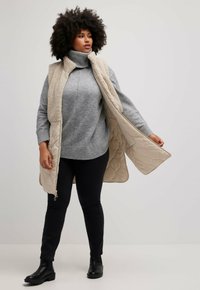 Langer beiger gesteppter Weste mit Kunstfellkragen über einem grauen Pullover und schwarzen Jeans, ergänzt durch schwarze Ankle Boots.