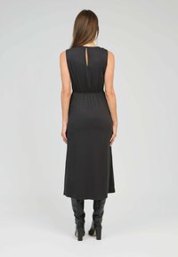 Robe noire sans manches avec une taille cintrée, présentant un détail en forme de trou de serrure à l'arrière et une jupe fluide. Associée à des bottes noires jusqu'aux genoux.