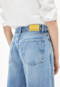 Hellblaue Denim-Jeans mit einem gelben Markenetikett, auf dem "ARMEDANGELS" steht. Merkmale sind zwei Gesäßtaschen und eine ausgebleichte Textur.