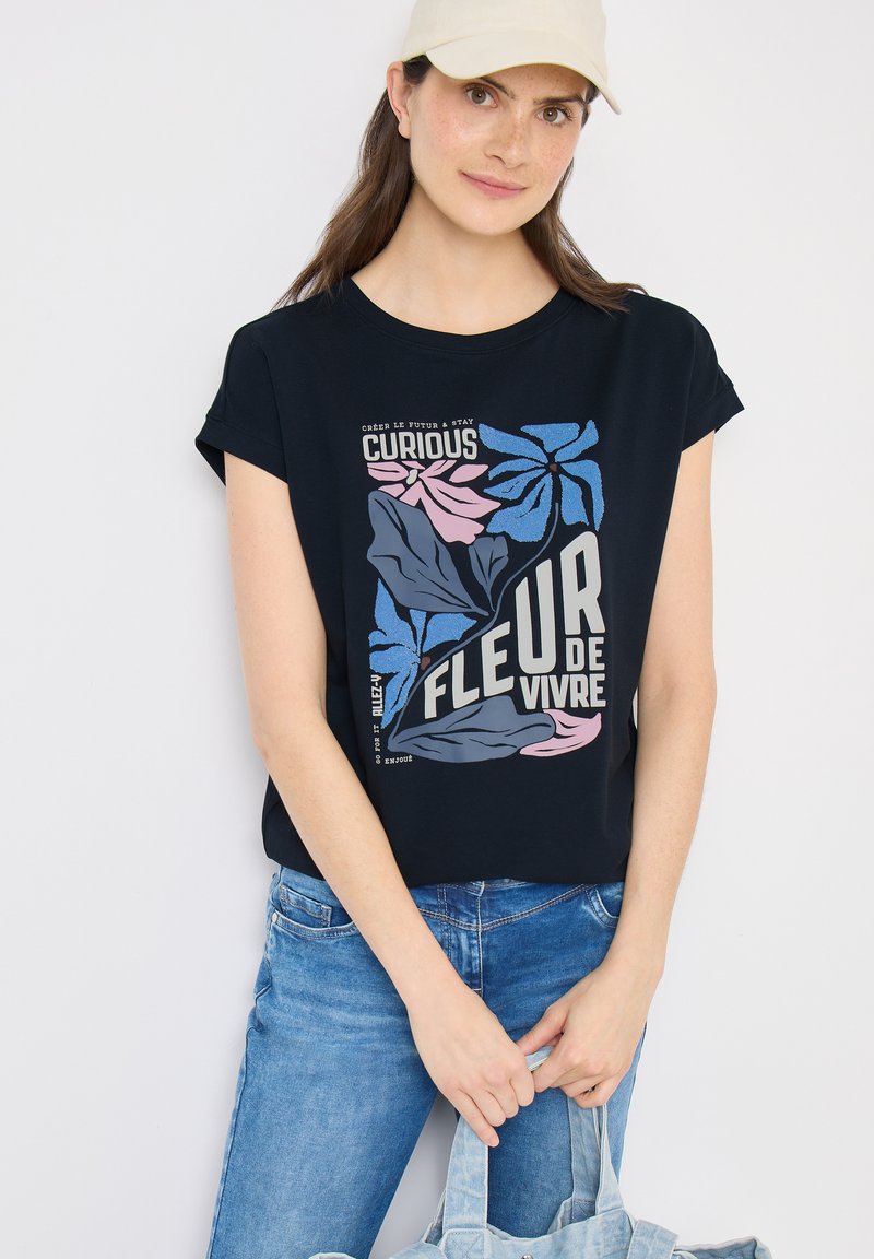 Femme portant une casquette beige, un t-shirt graphique noir avec un imprimé floral bleu et rose et du texte en français, un jean bleu, tenant un sac fourre-tout bleu clair.