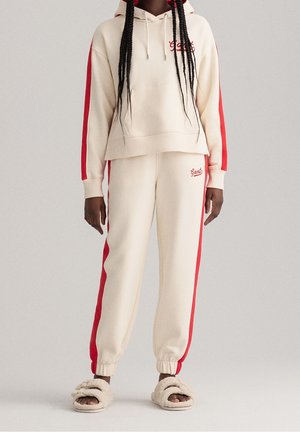 Personne portant un sweat à capuche couleur crème et un pantalon de jogging avec des bandes rouges, assortis à des chaussons crème duveteux, debout devant un fond uni.