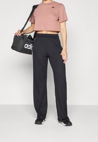 Svarta träningsbyxor med vita sidostrick, tillsammans med en ljusrosa kortärmad t-shirt med en liten logotyp, hålls av en person. Svart gymväska.