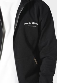 Felpa nera con zip realizzata in tessuto morbido, dotata di tasche frontali, cappuccio con coulisse e logo bianco sul petto, con un piccolo etichetta sul lato.