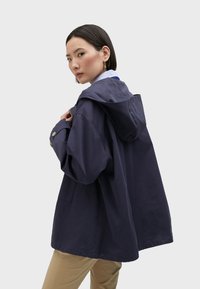 Navy blue hooded jacket made from cotton with a relaxed fit, buttoned cuffs, and a front pocket. Textured fabric with a matte finish.