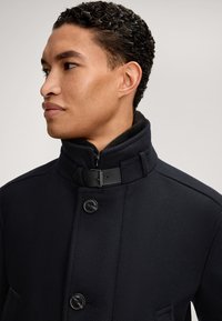 Cappotto blu navy con colletto alto, tasche laterali, chiusura con bottone e tessuto testurizzato. Presenta un'accento in fibbia nera sul colletto.