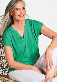 Groene v-hals korte mouw top gemaakt van zacht materiaal. Gecombineerd met witte relaxte broek. De achtergrond toont een geweven stoel.