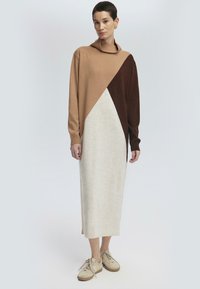 Femme portant une robe pull longue bicolore en beige, marron et crème, associée à des baskets décontractées beige, se tenant devant un fond uni.
