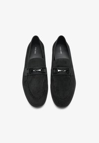 Schwarze Loafers aus strukturiertem Stoff mit gestreiftem Muster, ausgestattet mit einem metallischen Akzentband über dem Obermaterial. Rundliche Zehenform.