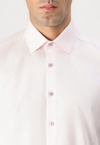 Helle rosa Bluse aus strukturiertem Stoff, mit spitzem Kragen und fünf passenden Knöpfen auf der Vorderseite. Keine weiteren Details oder Muster sichtbar.