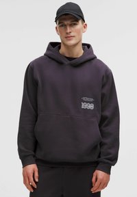 Lilla hættetrøje med kængurulomme og hvid tekstlogo på brystet. Fremstillet af blødt stof med ribbede manchetter og kant.