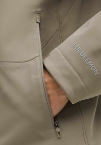 Chaqueta de color topo claro con tejido suave y texturizado. Cuenta con un bolsillo lateral con cremallera y el logo de 'Lululemon' en gris claro en la manga.