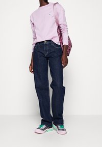 Camisa de manga larga de color púrpura claro, vaqueros rectos de color azul oscuro y zapatillas multicolor. La bolsa tiene un patrón de camuflaje en rosa.
