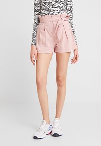 Shorts roses taille haute avec une ceinture nouée, des plis à l'avant et un tissu texturé. Associés à des baskets blanches et un haut à rayures noir et blanc.