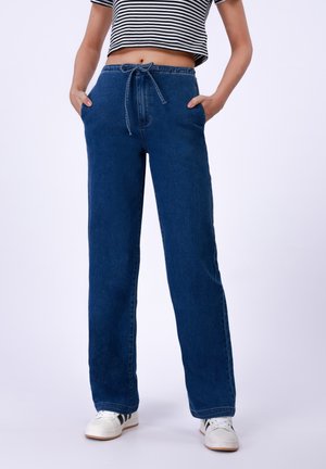 Pantaloni in denim blu con vita regolabile tramite cordoncino, tasche laterali, vestibilità ampia e gambe dritte. Indossati con sneakers bianche.