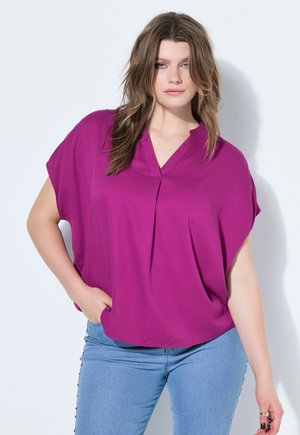 Młoda kobieta z luźnymi falującymi włosami, ubrana w jasną bluzkę z dekoltem w serek w kolorze magenty oraz jasnoniebieskie jeansy z ozdobnymi ćwiekami po bokach.