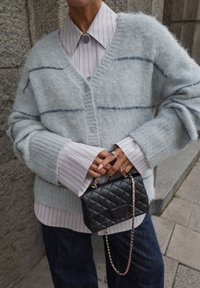 Grau-mohair Cardigan mit blauen Streifen, über einem hellgestreiften Shirt getragen. Schwarze, gesteppte Umhängetasche mit Kettenriemen. Dunkle Jeans.