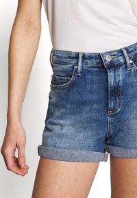 Shorts de denim en un lavado azul desgastado con dobladillos enrollados, que presentan un clásico diseño de cinco bolsillos y cierre de botón metálico en la cintura.