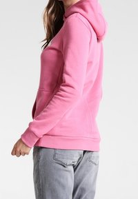 Sudadera de felpa rosa con bolsillo frontal y capucha con cordón. Presenta puños y dobladillo acanalados, textura suave y un ajuste ligeramente holgado.
