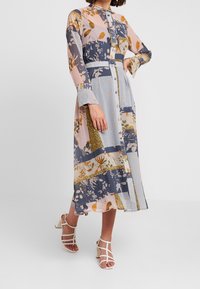 Robe longue boutonnée avec un mélange de motifs floraux et géométriques dans des couleurs pastel, à manches longues et à taille froncée. Associée à des sandales à lanières.