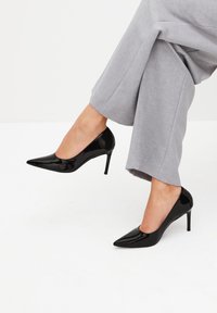 Décolleté con tacco alto in vernice nera con punta affilata e silhouette elegante, abbinate a pantaloni grigi a gamba larga.