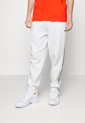 Mand iført hvide Lacoste joggingbukser med sidelogo-bånd, hvide sneakers og en orange skjorte, stående mod en ensfarvet lys baggrund.