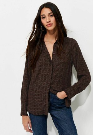 Woman with long dark hair wearing hoop earrings, a dark patterned button-up shirt, and blue jeans, posing with one hand in pocket.