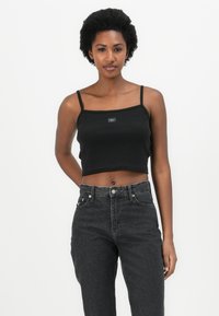 Black ribbed crop top with thin straps and a rectangular logo patch, paired with high-waisted dark denim jeans.