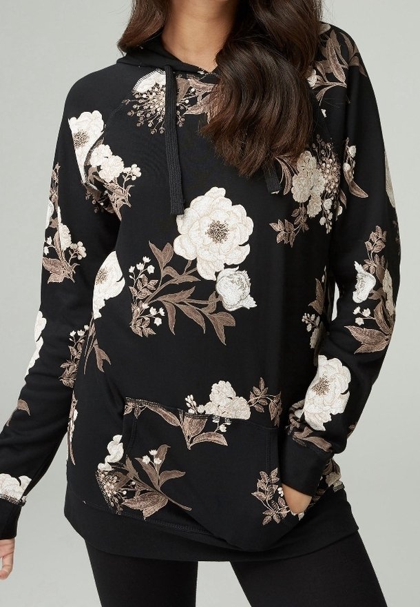 Sweat à capuche noir avec motifs floraux blancs et beiges porté par une personne aux longs cheveux bruns, mains partiellement dans la poche avant.