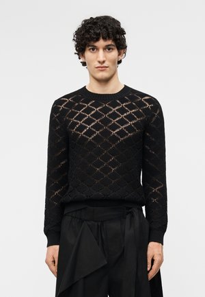 Jeune homme aux cheveux courts et bouclés portant un pull en maille noir à manches longues avec un motif en losange et un pantalon noir ample, sur un fond uni.