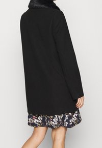 Cappotto nero in lana con colletto in pelliccia sintetica, taglio dritto, maniche lunghe e tasche laterali; indossato sopra una gonna a balze con stampa floreale.