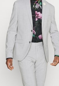 Uomo che indossa un completo grigio chiaro con due bottoni e una camicia floreale nera con motivi di fiori rosa e verdi, mani rilassate ai lati.