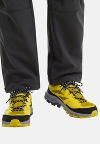 Scarpe da trekking gialle e nere con lacci bianco e nero indossate con pantaloni da esterno neri su uno sfondo neutro.