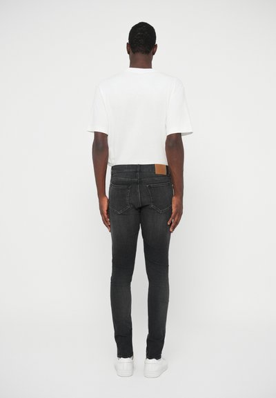 Jeans negre slim-fit cu buzunare la spate, petic din piele maro la talie, asortați cu un tricou alb simplu și adidași albi.