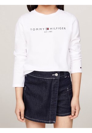 Kind trägt weißes Langarmshirt mit "Tommy Hilfiger Est. 1985" Logo und asymmetrischen dunklen Denimrock.