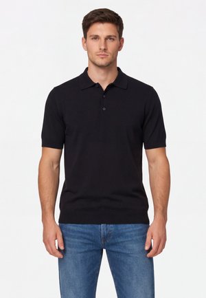 Uomo che indossa una polo nera a maniche corte semplice e jeans blu, in piedi davanti a uno sfondo bianco uniforme.