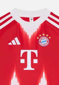 Maglia sportiva rossa con colletto bianco e tre strisce. Presenta un grande logo bianco a "T" e lo scudetto del FC Bayern München con motivo a quadri blu.