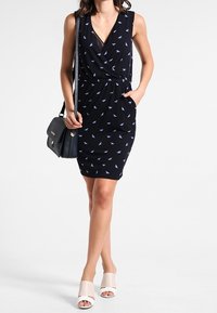 Czarna sukienka bez rękawów z fasonem kopertowym i małymi wzorami niebieskich ptaków, z elastyczną talią i bocznymi kieszeniami. Zestawiona z torebką.